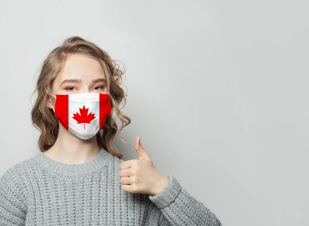 Happy woman in face mask holding thumb up with national flag Canada background. Flu epidemic and virus protection concept