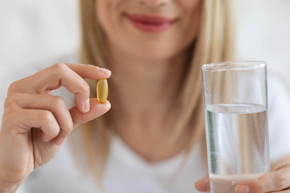 Cropped of blonde lady holding pill yellow gelatin capsule and glass of water, unrecognizable lady taking supplement vitamin D after waking up in the morning, blurred background