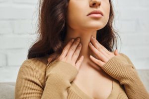 Cropped view of young brunette woman in brown jumper checking thyroid gland on neck while sitting